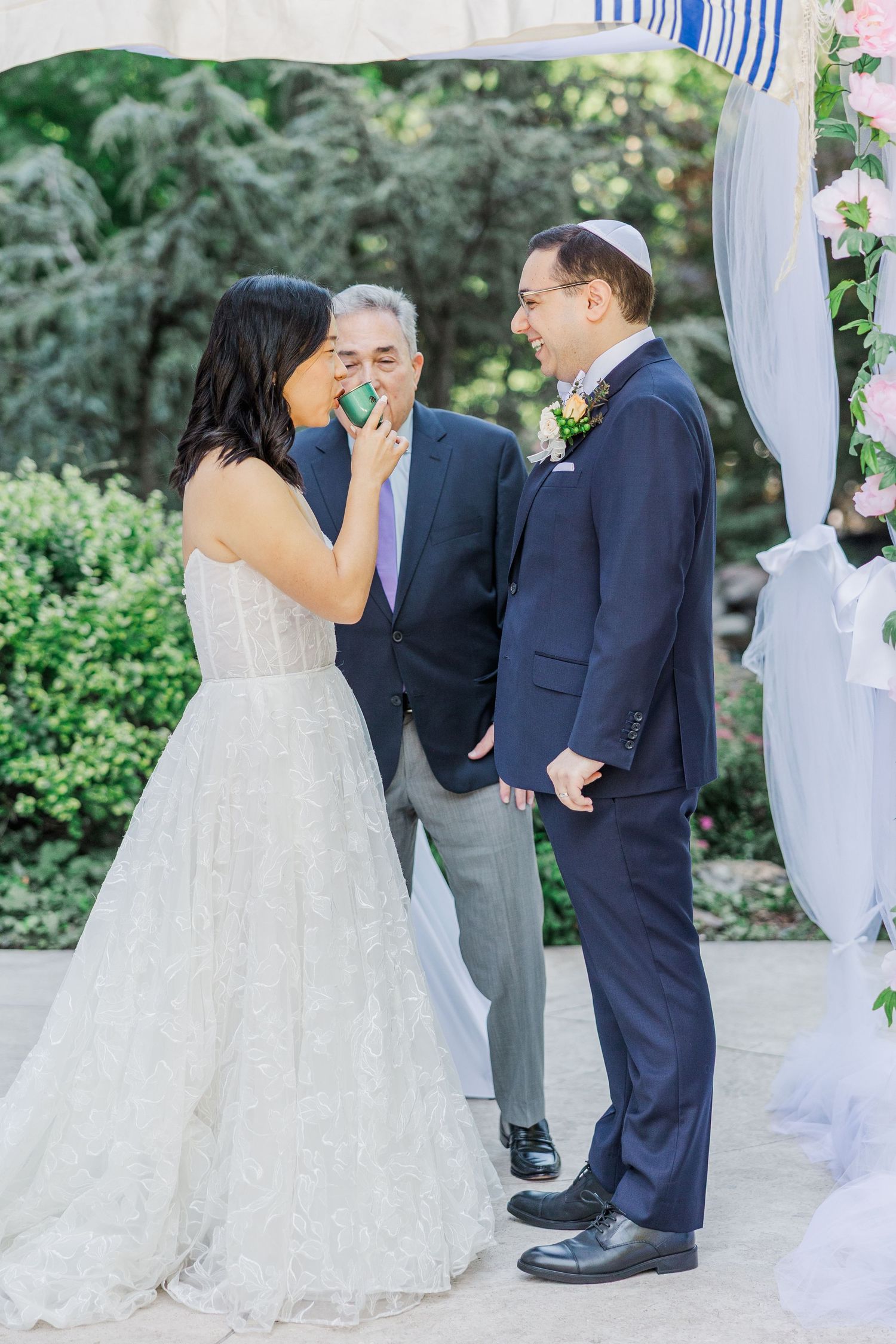 Bride and groom share a joyful moment during outdoor Jewish wedding ceremony under floral chuppah.