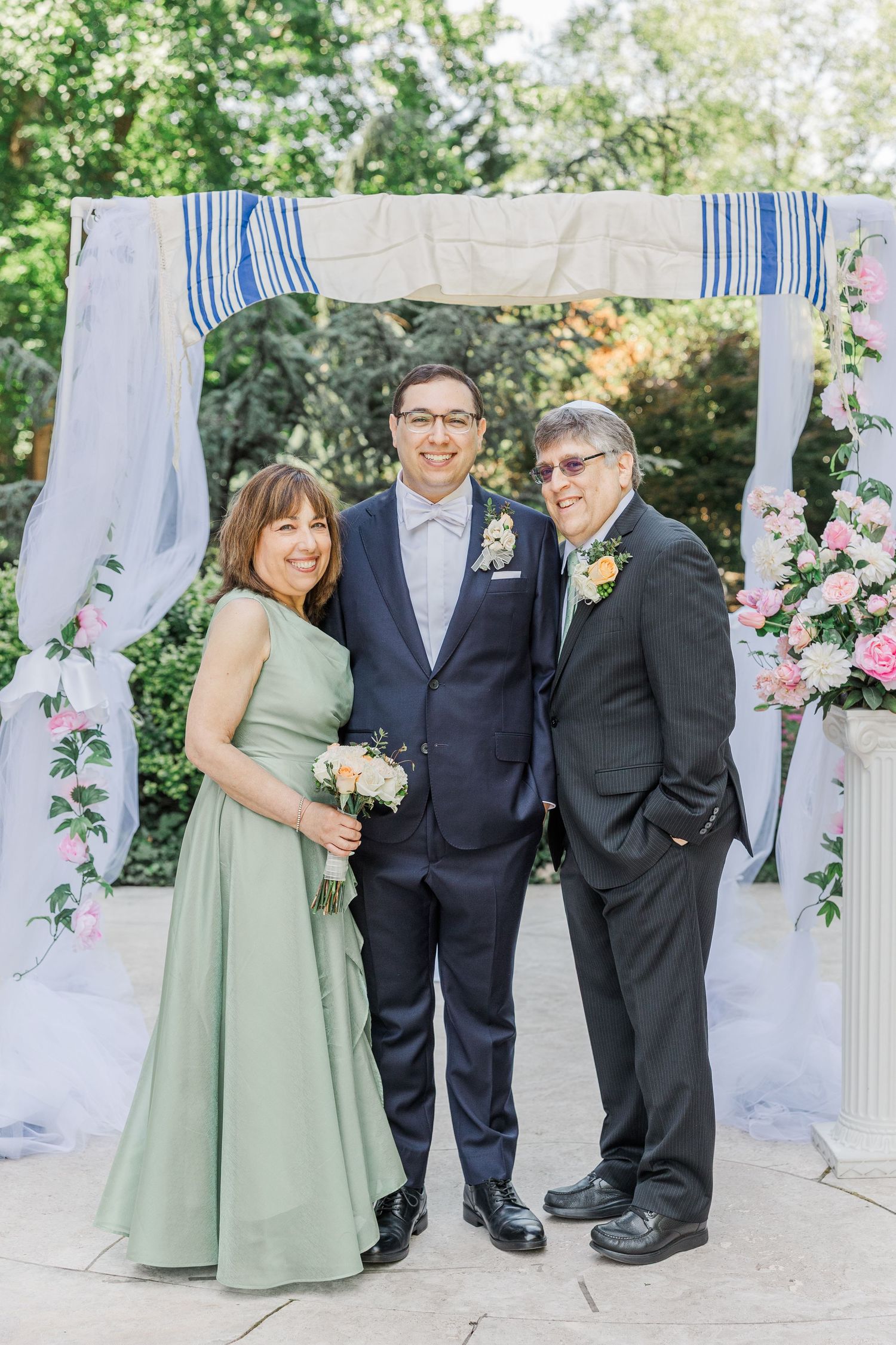 Groom in navy suit poses with parents under floral chuppah at outdoor wedding ceremony.