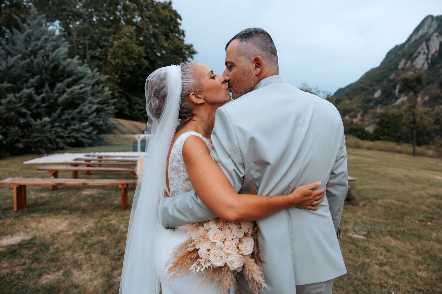 Bride and groom sharing a kiss outdoors at a wedding ceremony with mountain scenery and wooden benches in the background.