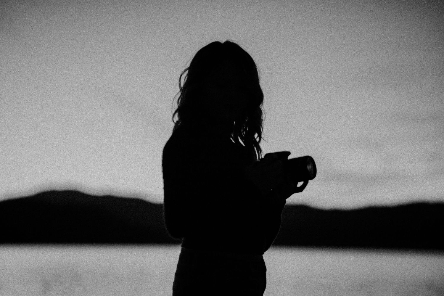 Silhouette of a woman holding a camera against a twilight sky with mountains and lake in the background.