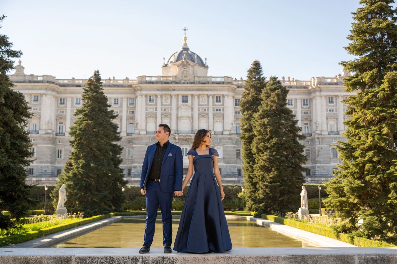 Couple in navy formal wear posing in front of the Royal Palace of Madrid gardens.
