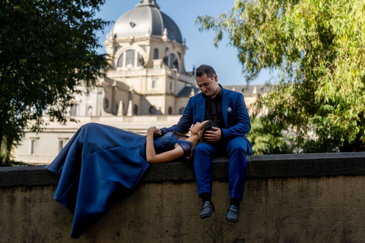 Couple in matching blue outfits posing on a stone wall with a domed building and trees in background.