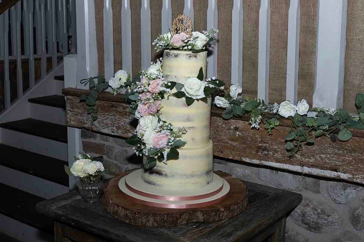 Three-tier semi-naked wedding cake adorned with pink and white roses and greenery, displayed on a wooden slice stand.