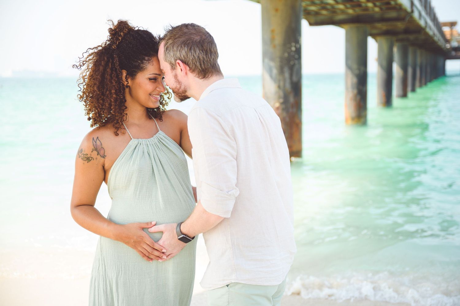 Pregnant couple embracing on tropical beach near pier, woman in mint dress with tattoo, man kissing her forehead.