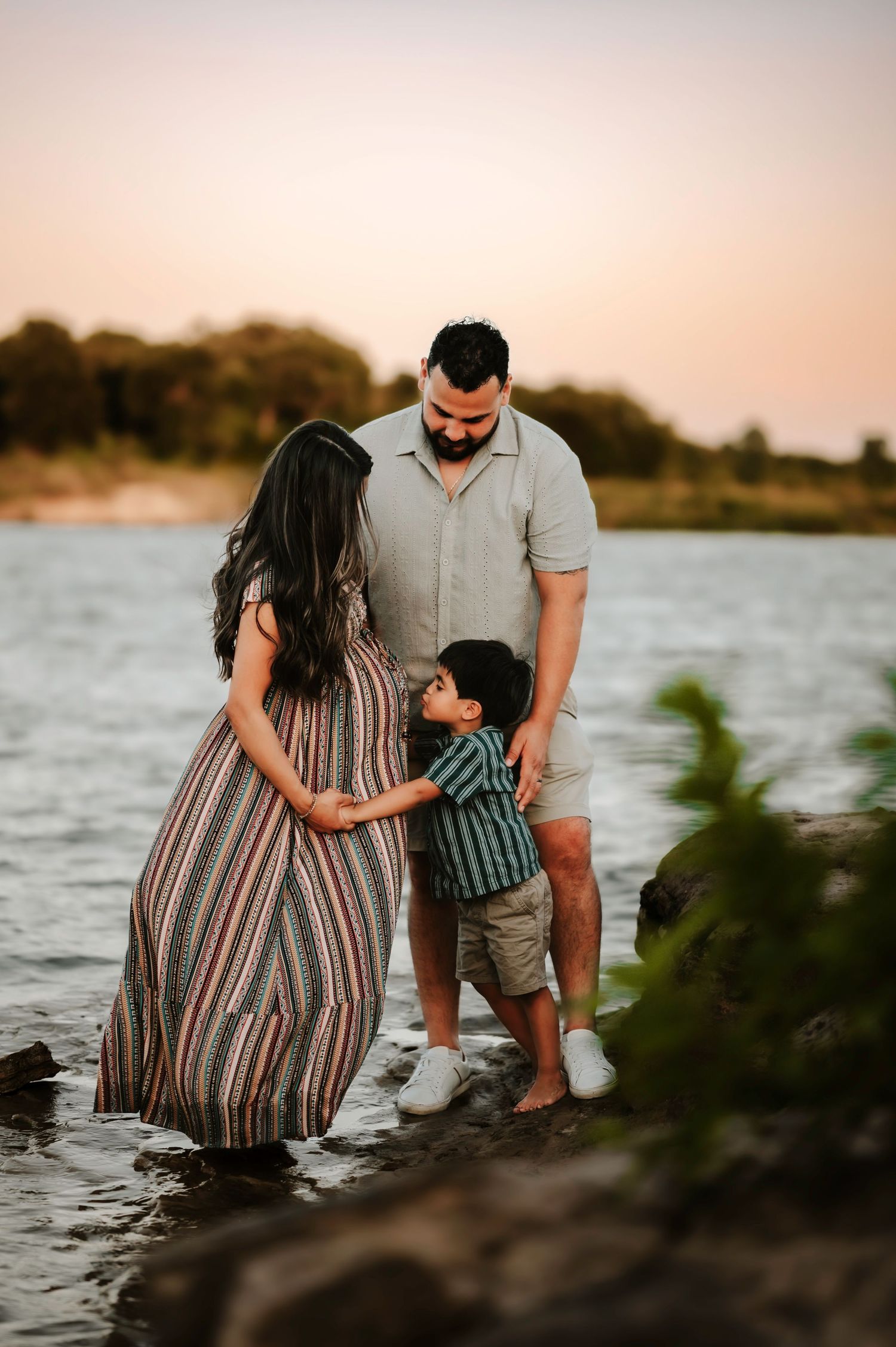 Pregnant woman in striped maxi dress with husband and son standing at waters edge at sunset.