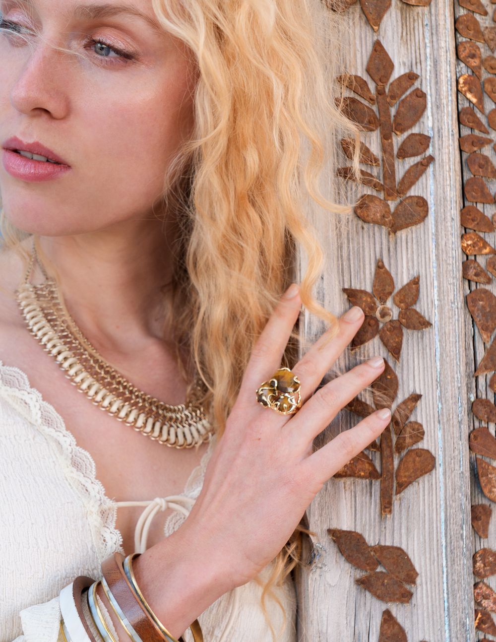 Blonde woman wearing gold layered necklace, floral ring, and silver bangles, posing against rustic wooden wall.