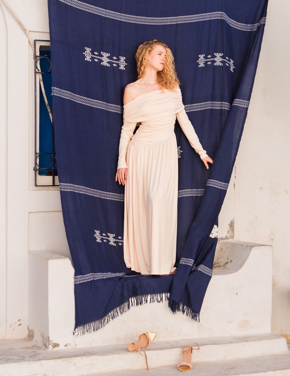 Woman in elegant off-shoulder cream pleated gown poses against navy blue woven textile backdrop outdoors.