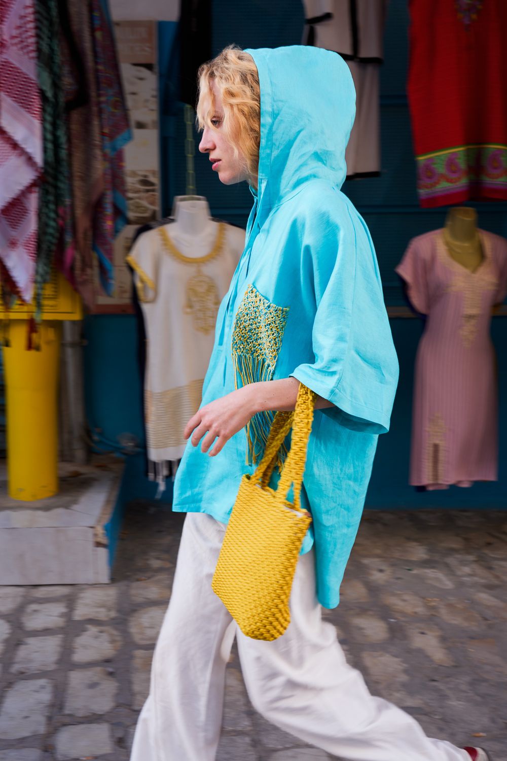 Woman in turquoise hooded top and white pants carries yellow crochet bag while walking past colorful clothing shop.