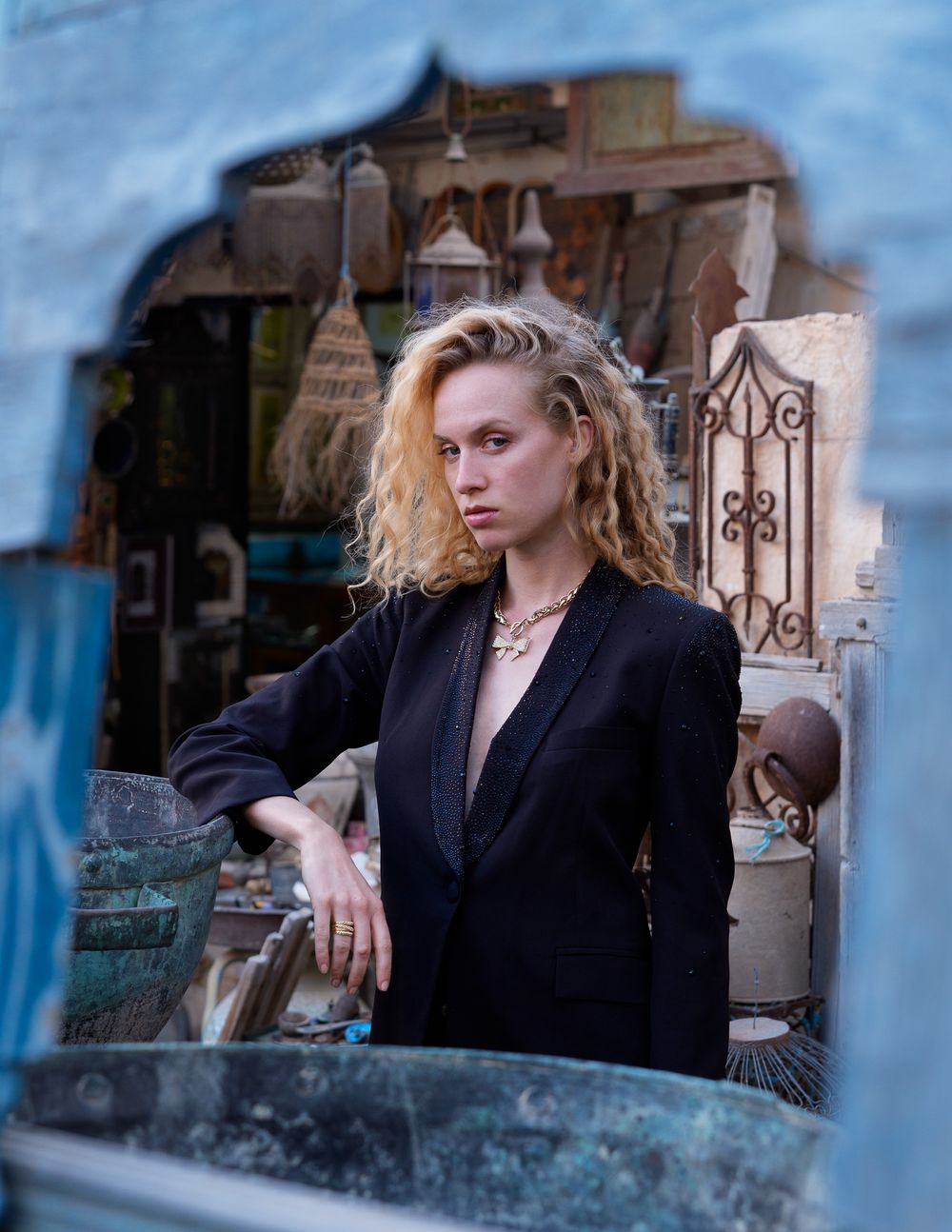 Blonde curly-haired woman in black blazer poses among antique decor in an outdoor market setting.
