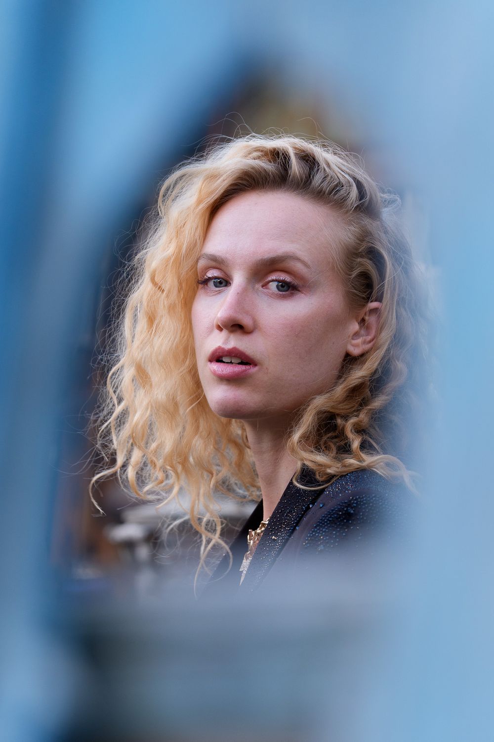 Young woman with curly blonde hair gazing thoughtfully, framed by a blurred blue background.