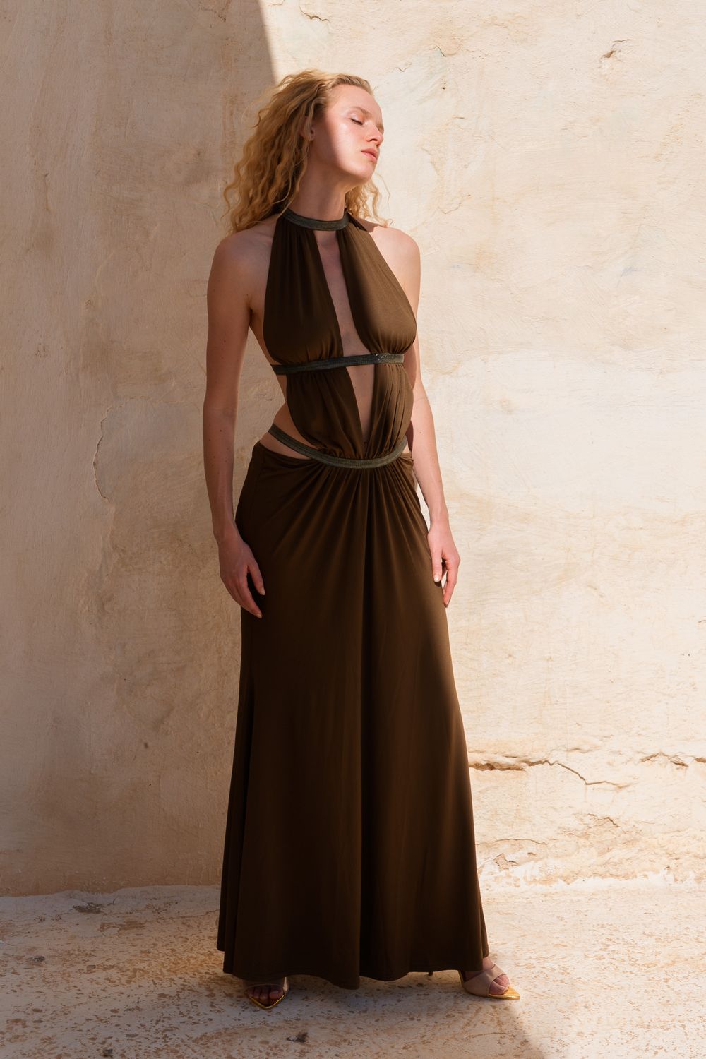 Woman wearing a dark olive halter maxi dress with cutout details, standing against a textured beige wall.