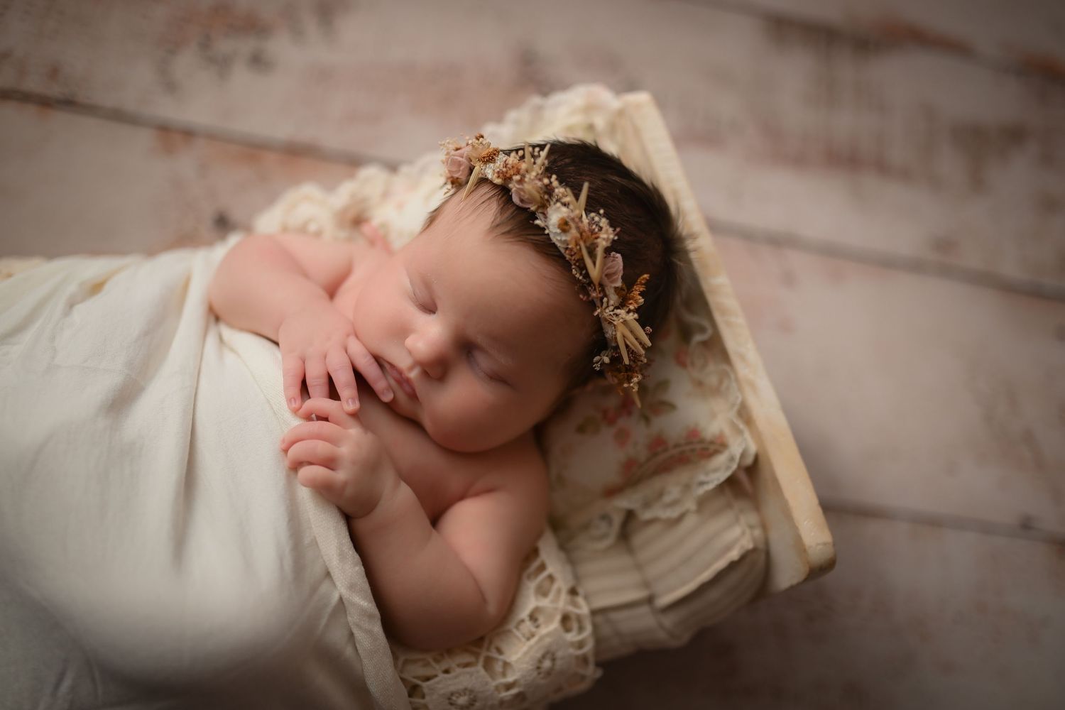 Sleeping newborn baby girl wrapped in cream lace with a gold floral headband in a rustic wooden crate.