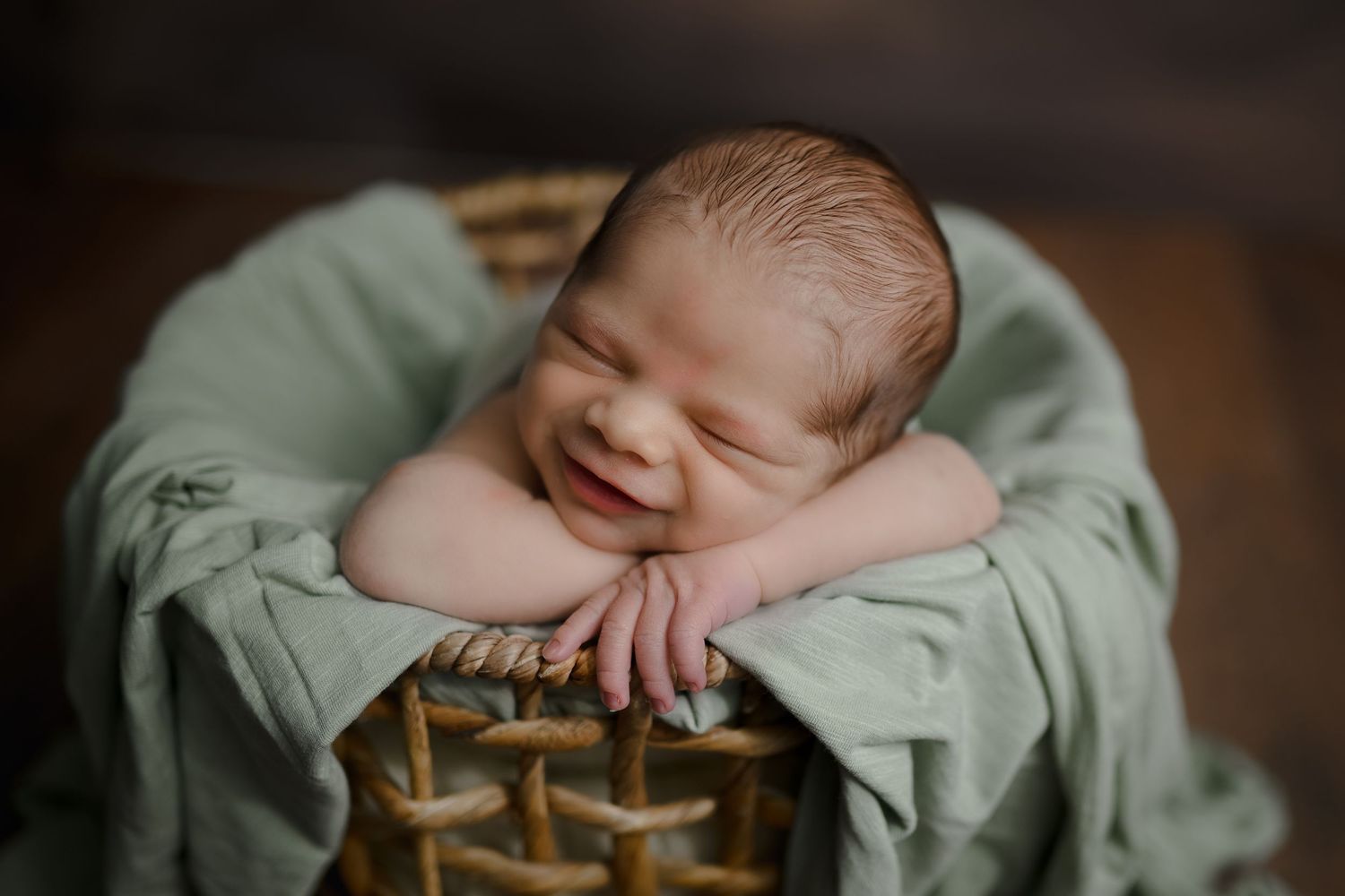 Smiling newborn baby sleeping in a wicker basket wrapped in a soft green blanket during a studio photo session.