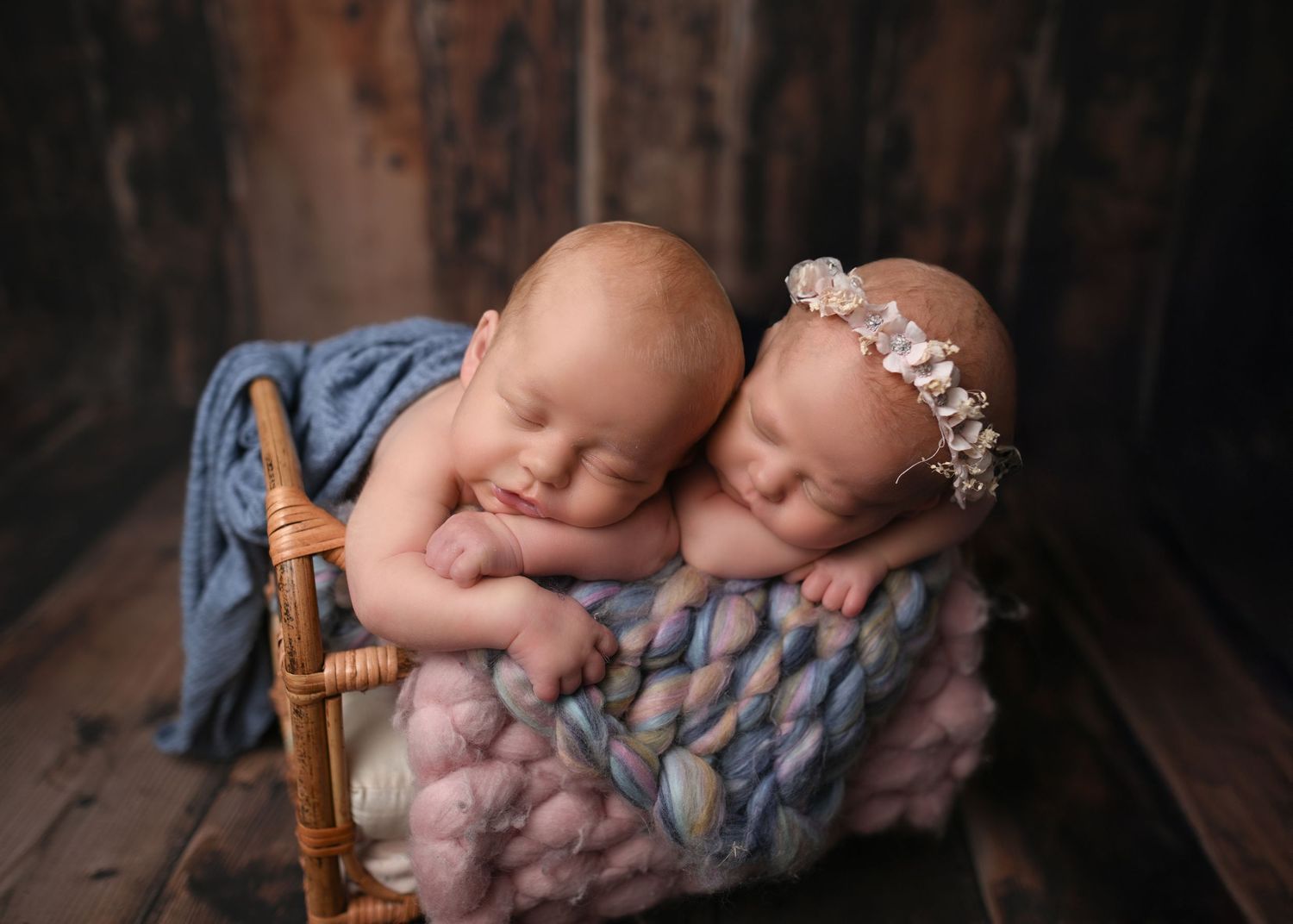 Two sleeping newborn twins nestled together in a rustic wooden basket with soft colorful knit blanket.