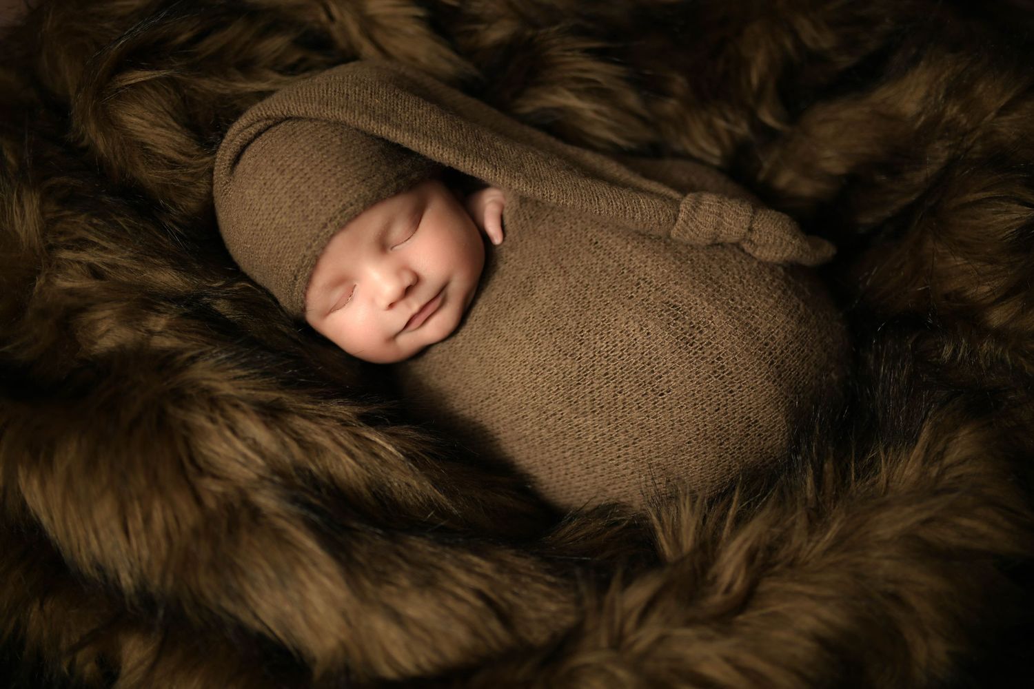 Newborn baby swaddled in brown knit wrap and bonnet, sleeping peacefully on a soft fur blanket.
