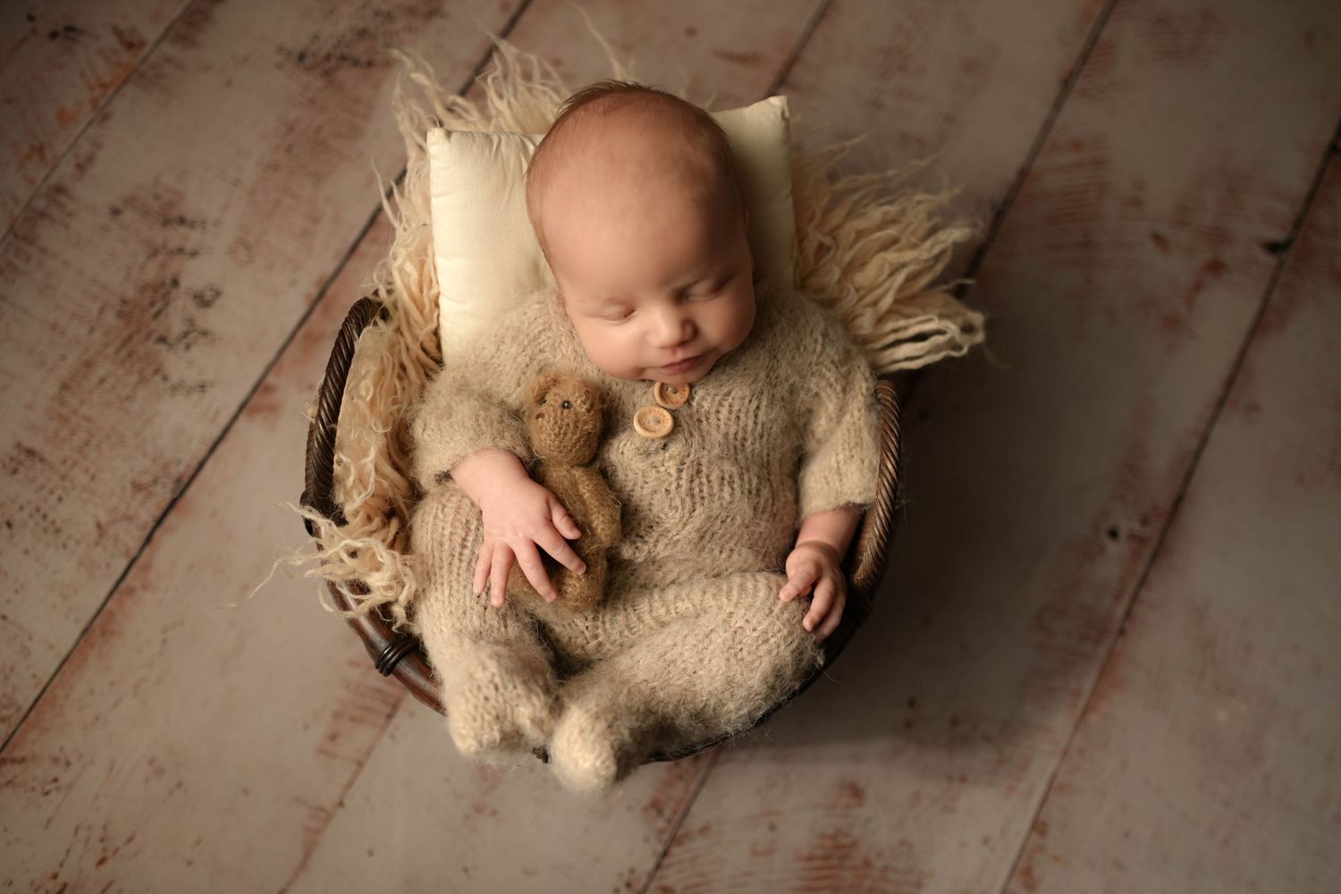 Newborn baby sleeping peacefully in a basket wearing a knit onesie, holding a tiny teddy bear.