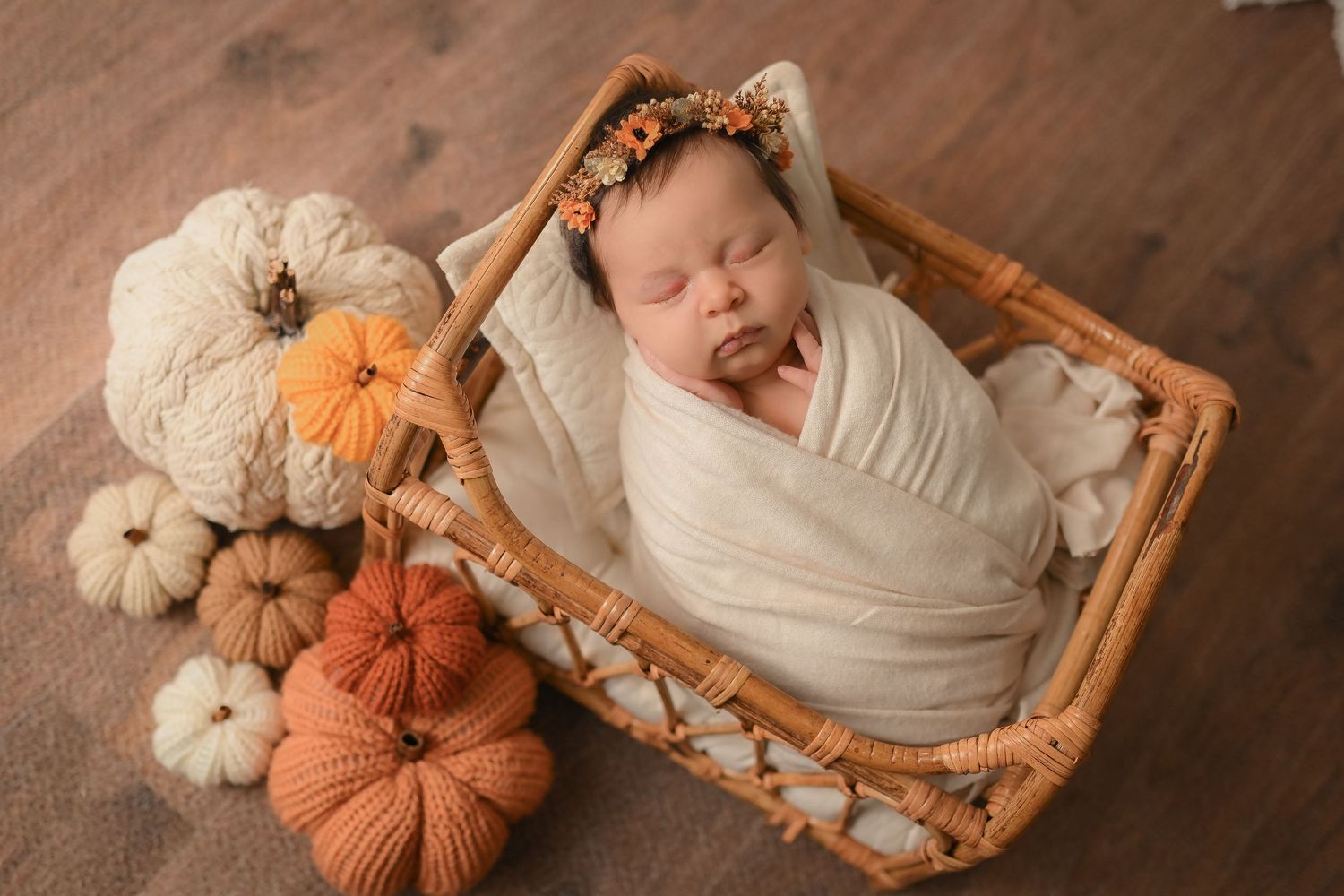 Newborn baby wrapped in cream swaddle sleeping in wicker basket with fall pumpkins and floral headband.