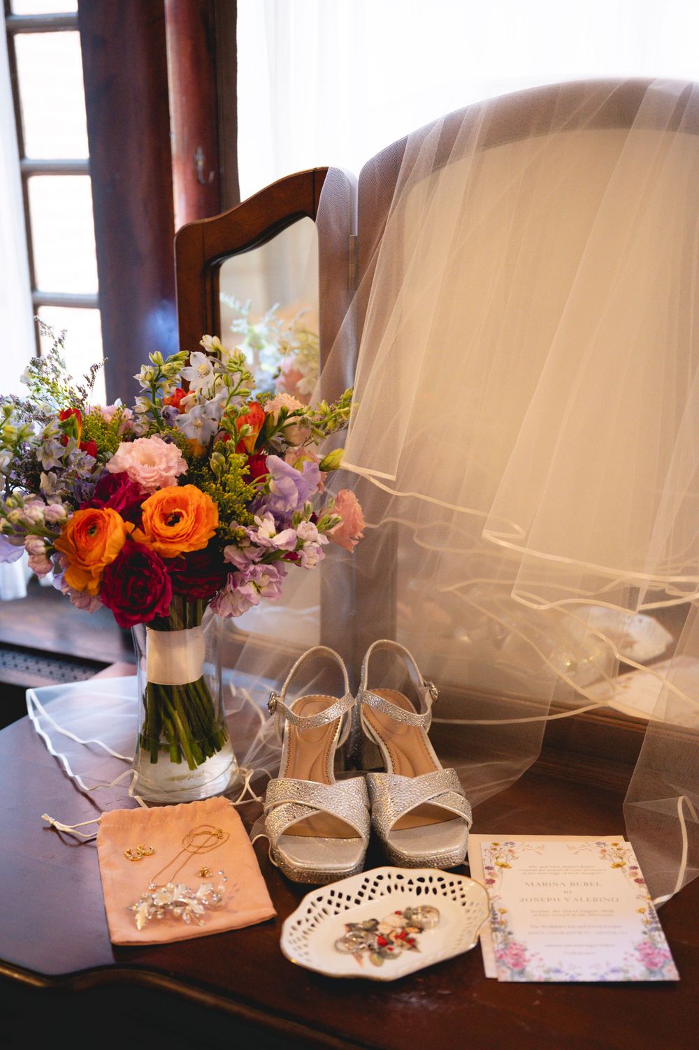 Bridal flat lay with colorful bouquet, white heels, veil, jewelry, and wedding invitation on a wooden table.