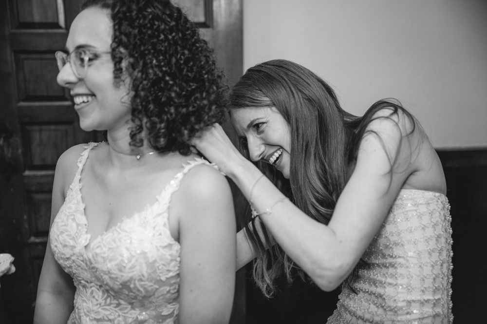 Black and white photo of two women laughing together, one in a wedding dress being helped by a smiling bridesmaid.