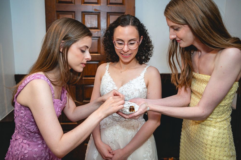 Bride in white lace gown shares a sweet moment with two bridesmaids examining a small treat.