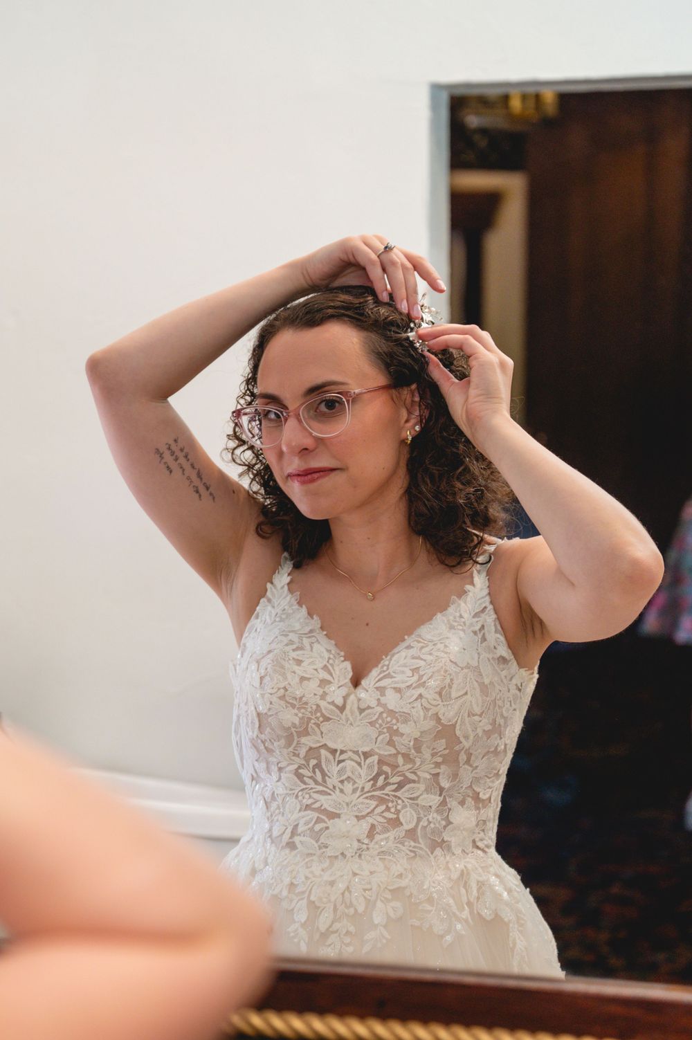 Bride in lace wedding dress and glasses adjusting her curly hair in a mirror before her wedding ceremony.