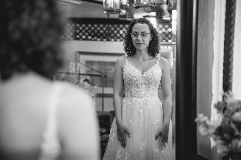 Black and white photo of a bride in a lace wedding gown gazing at her reflection in a mirror.