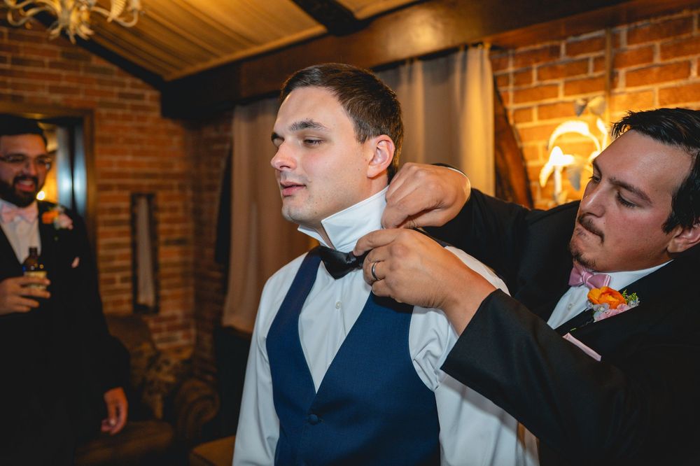 Groomsman adjusting bow tie on groom in vest during wedding preparation indoors.