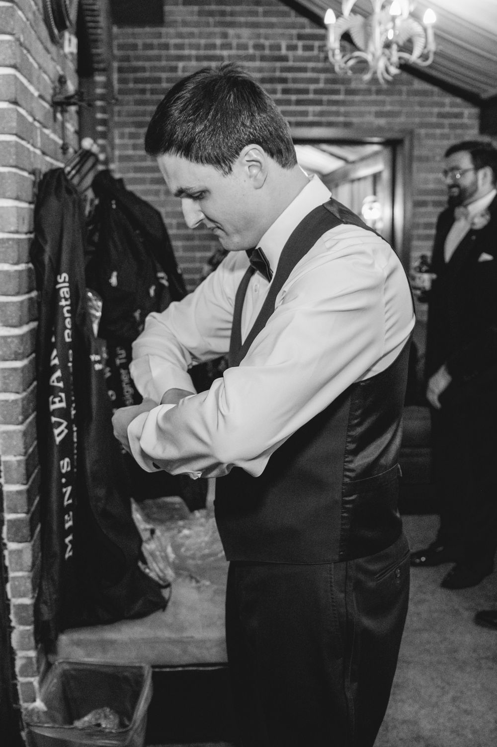Groom in vest and bow tie adjusting cufflinks before wedding ceremony in black and white photo.
