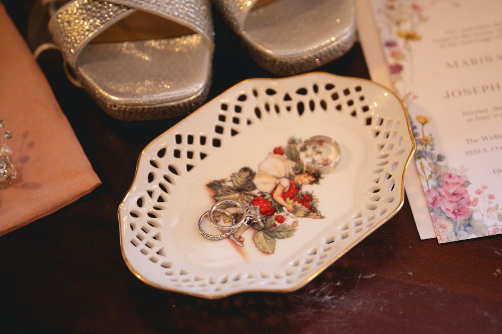 Vintage porcelain trinket dish with cherub design holding jewelry, beside silver heels and wedding invitation.