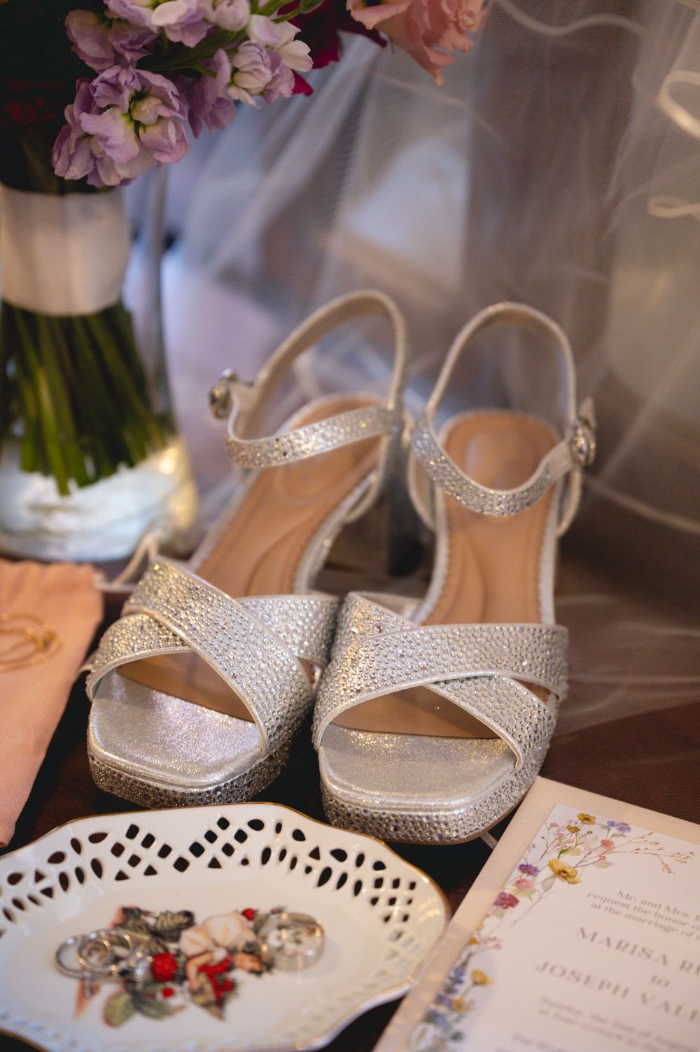 Silver glitter wedding heels displayed beside floral bouquet, jewelry dish, and wedding invitation on dark surface.