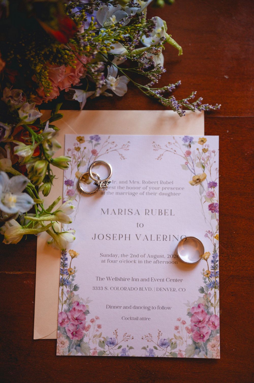 Wedding invitation for Marisa Rubel and Joseph Valerino with gold rings and floral bouquet on wooden table.