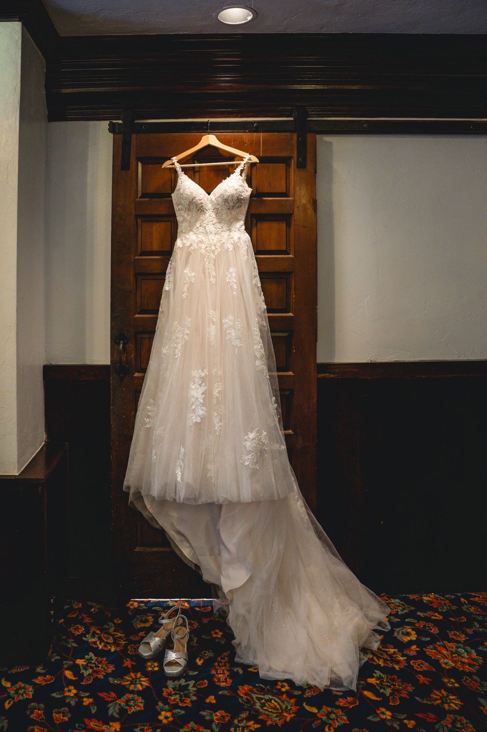 Elegant lace A-line wedding dress with floral appliques hanging on wooden door with bridal heels below.