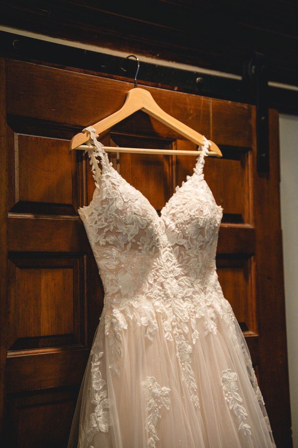 Elegant lace A-line wedding dress with V-neckline hanging on wooden hanger against rustic wood door.