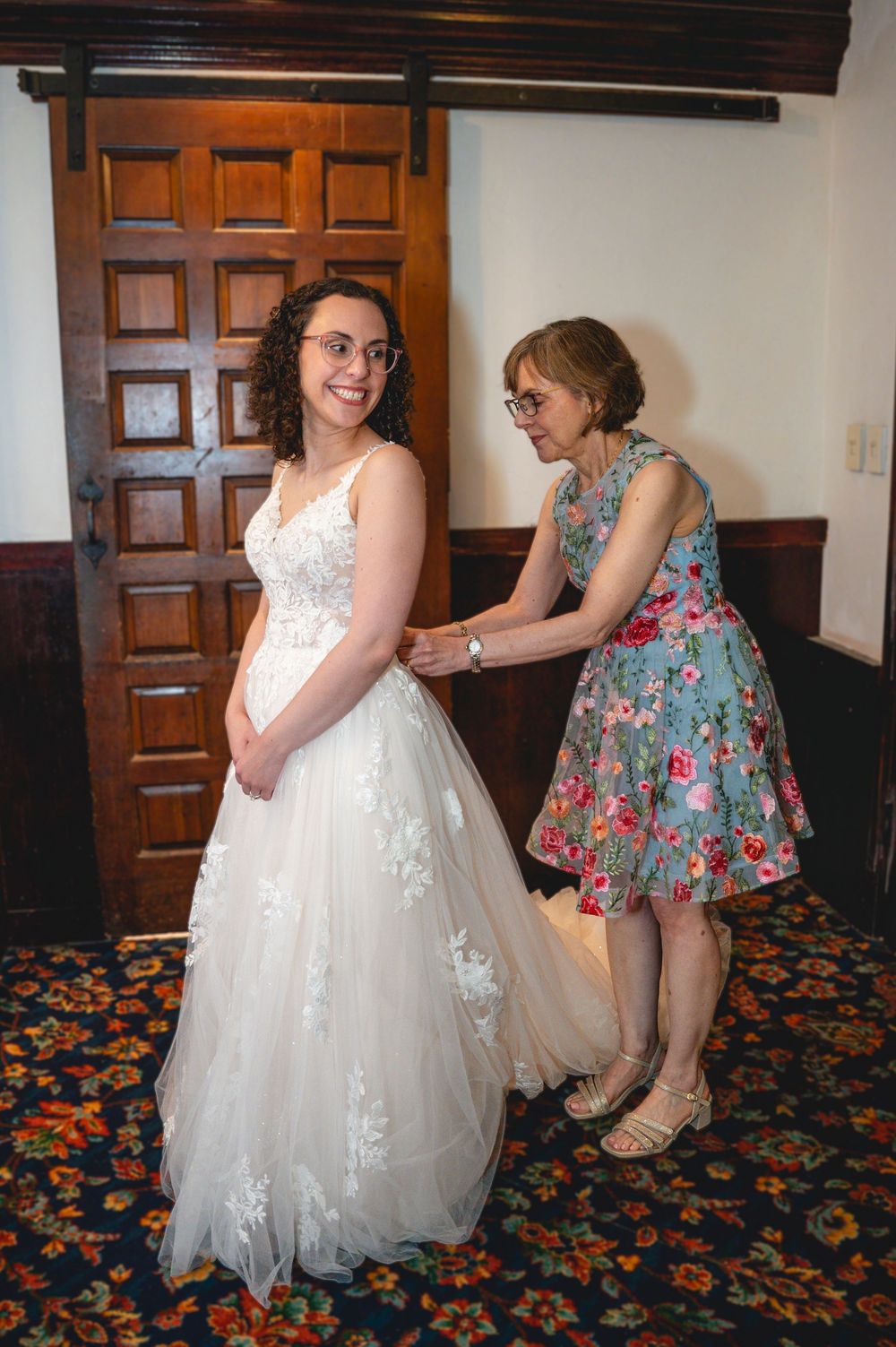 Bride in white lace gown smiling as woman in floral dress helps fasten her wedding dress in elegant wood-paneled room.