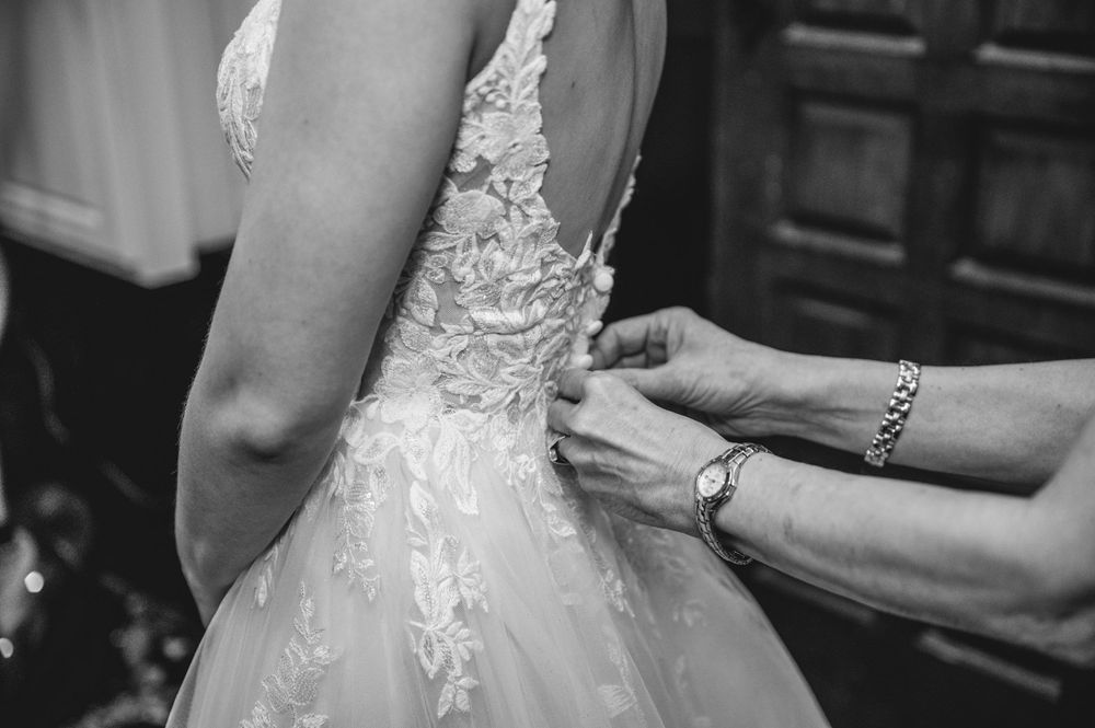 Black and white photo of hands fastening the back of a lace wedding dress on a bride.