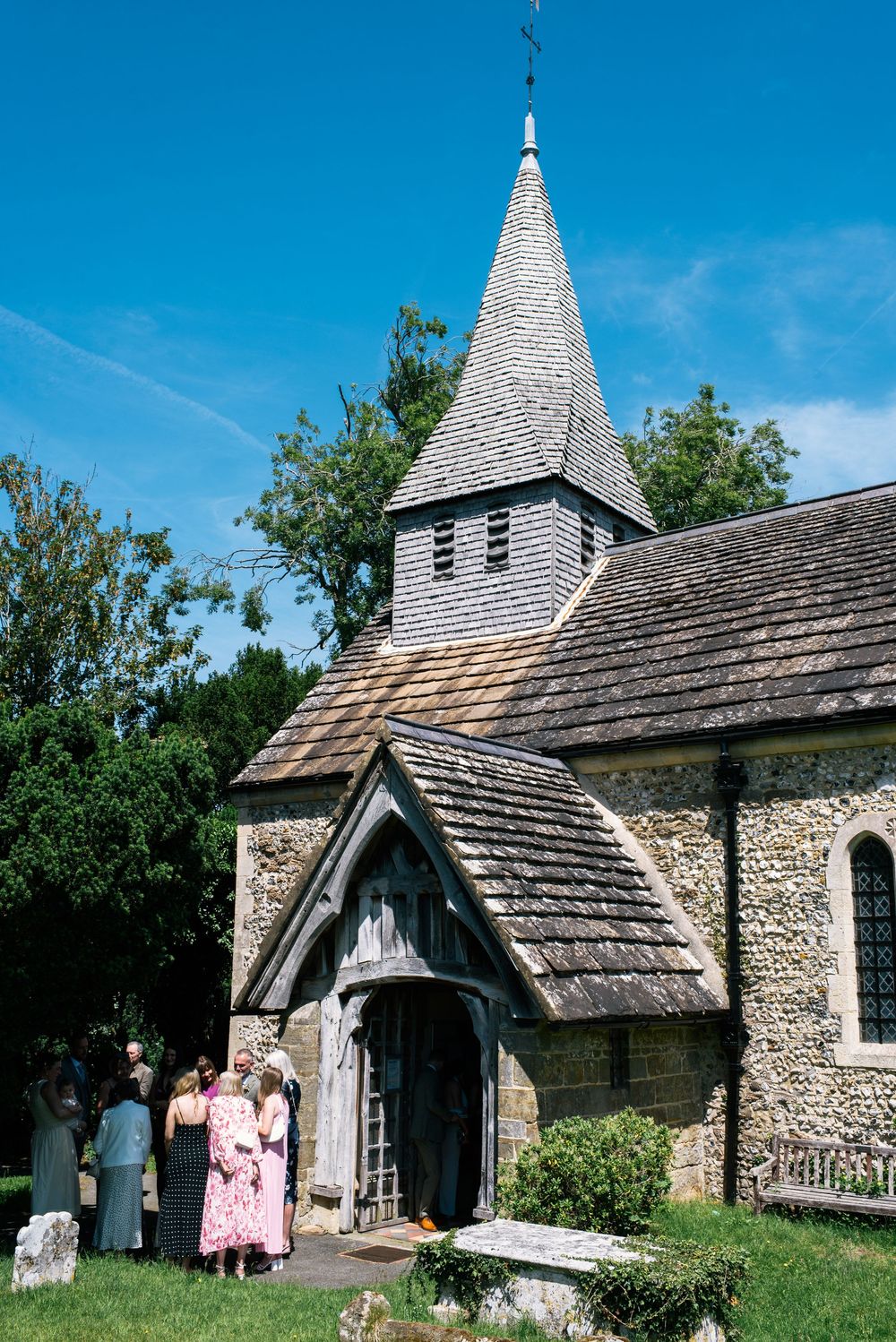 A charming stone English country church with a pointed spire under a bright blue sky with wedding guests gathered at the entrance.