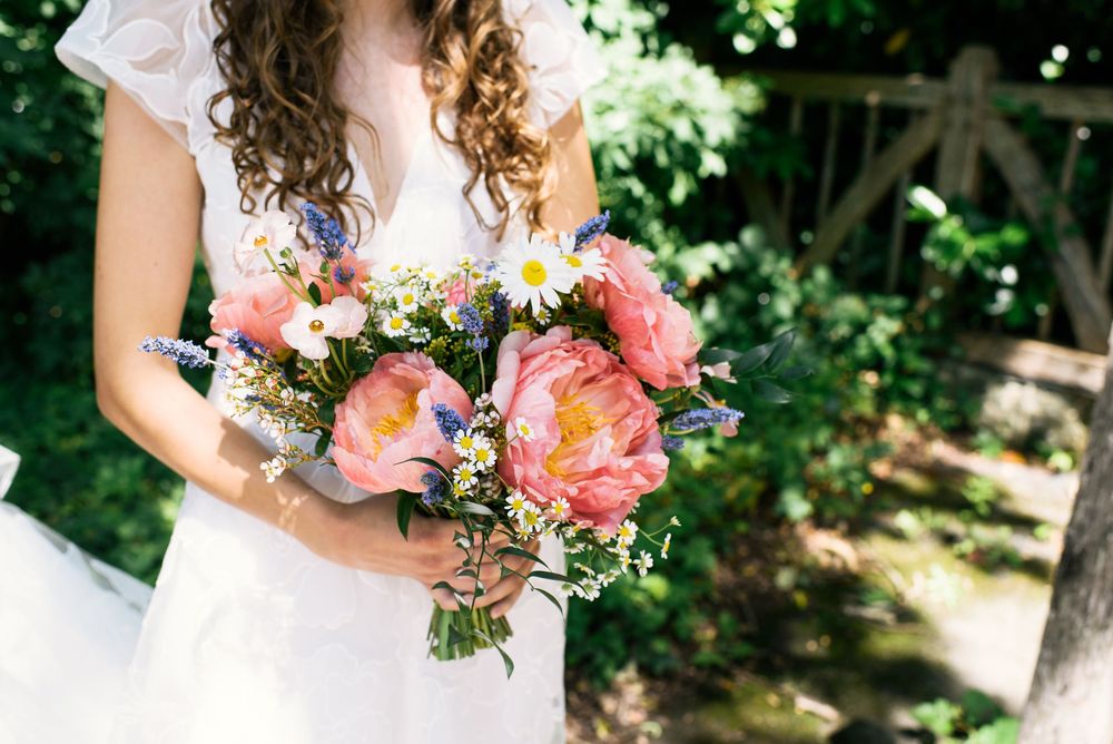 Bride in white dress holding a vibrant wedding bouquet with pink peonies, daisies, and wildflowers outdoors.