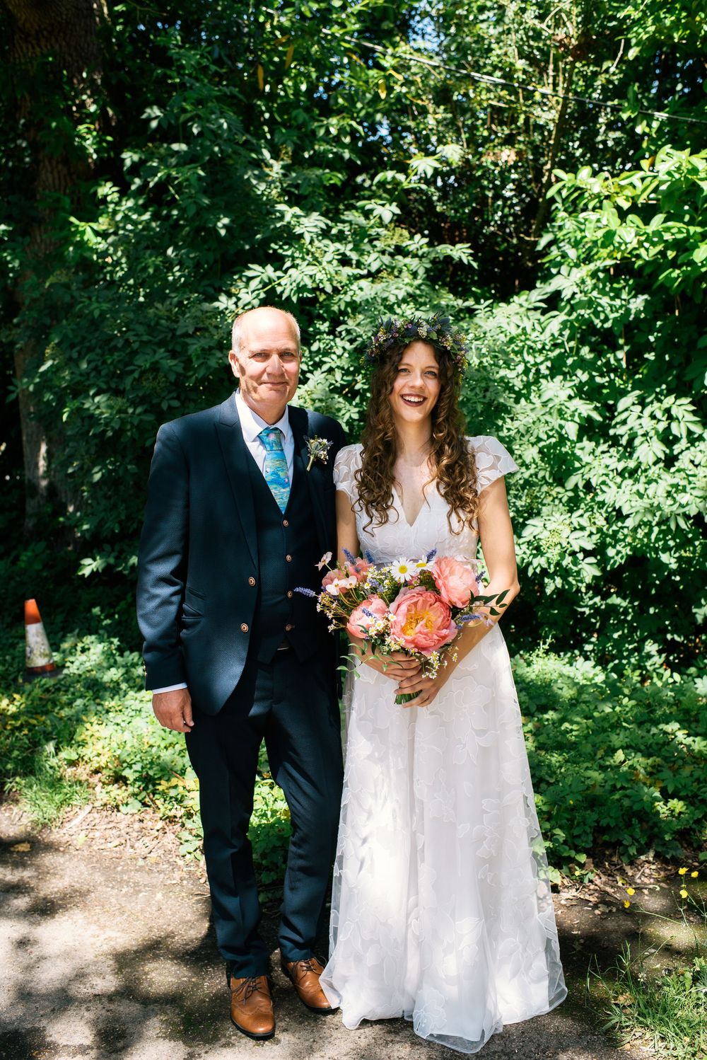 Bride in lace gown holding pink bouquet stands with groom in navy suit outdoors amid lush green trees.