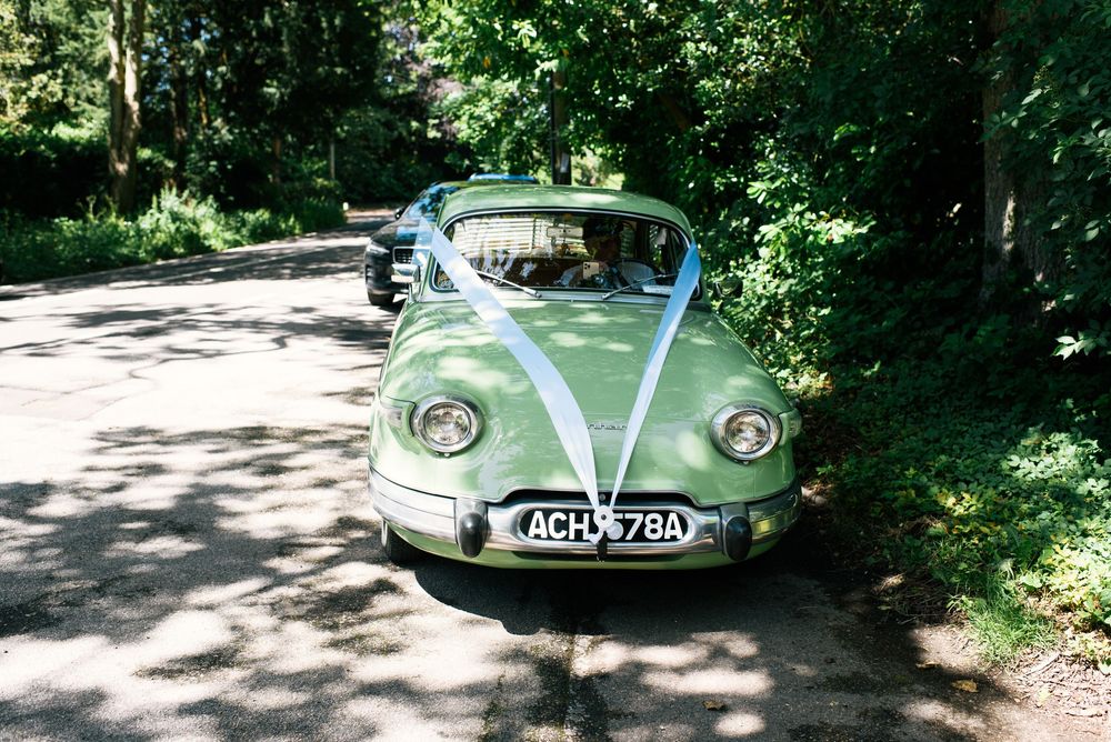 Vintage green wedding car decorated with white ribbons on a tree-lined road.