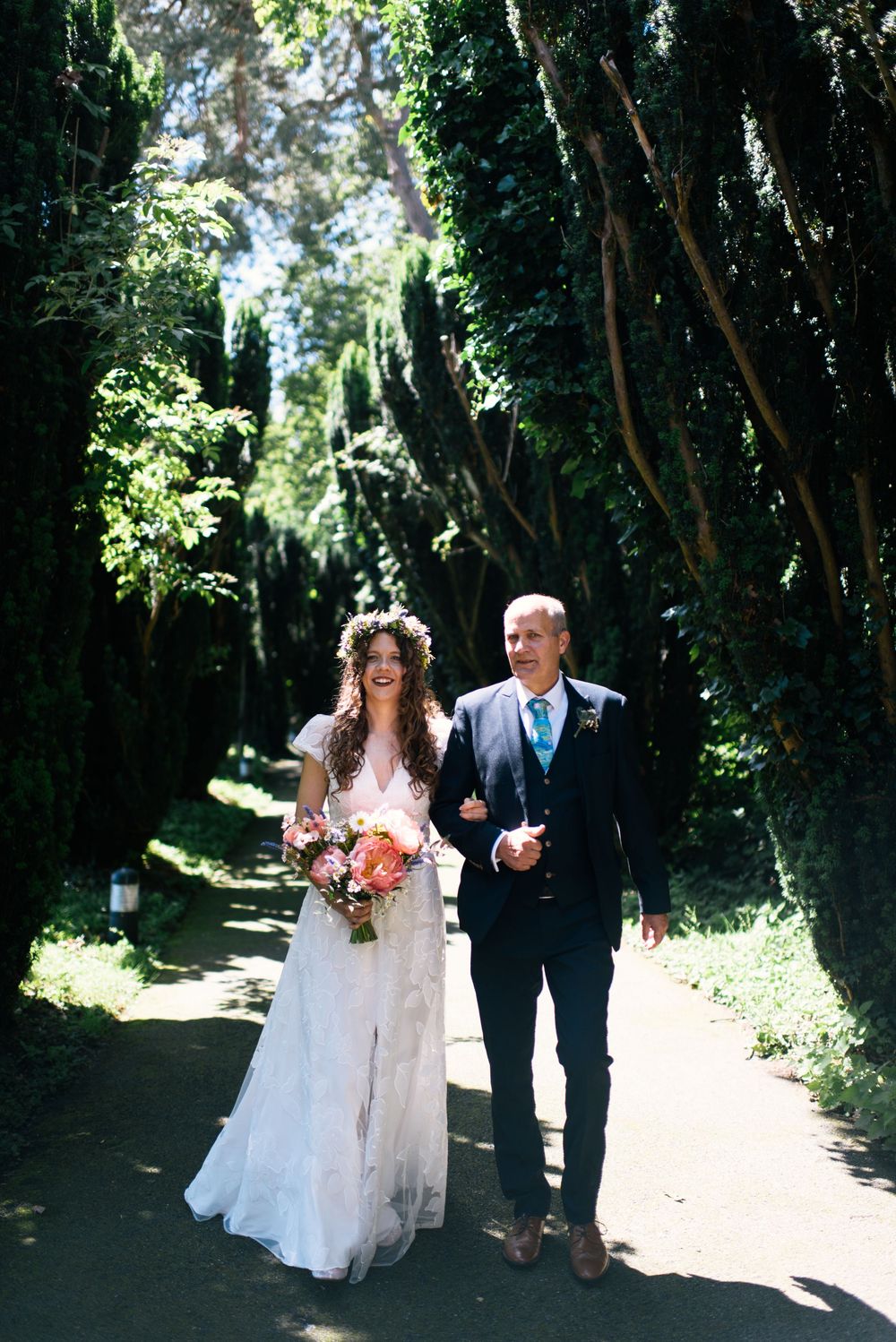 Bride in white lace gown and floral crown walks with escort down tree-lined path holding colorful bouquet.