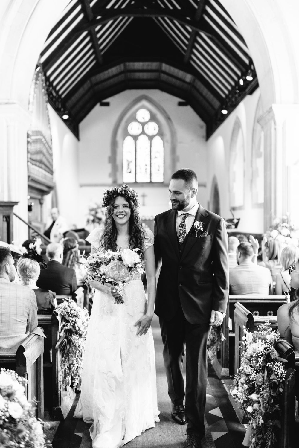 Black and white photo of a smiling bride in floral crown and groom walking down the church aisle after their wedding ceremony.