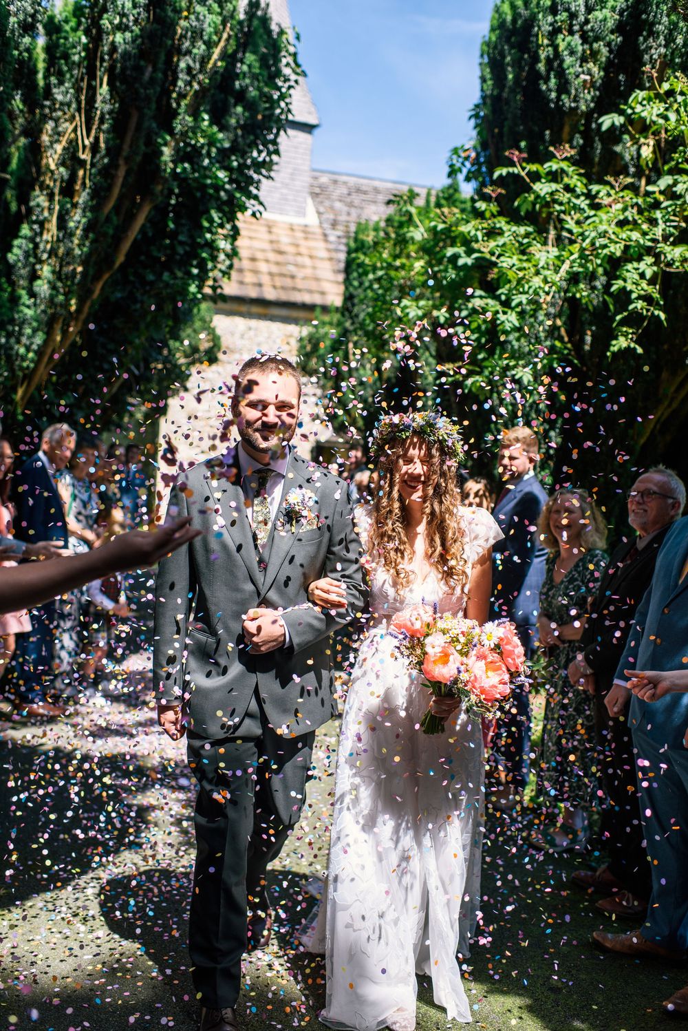 Joyful newlyweds walk through a shower of colorful confetti thrown by guests outside a historic stone church on a sunny day.