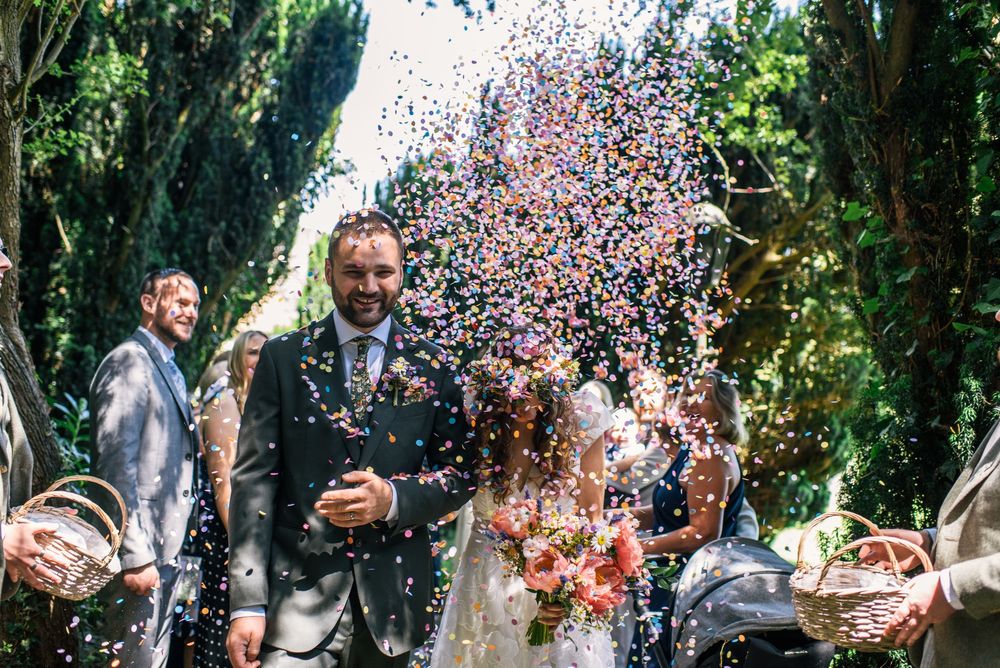 Joyful newlyweds walk through colorful confetti shower outdoors, surrounded by wedding guests holding baskets in lush garden setting.