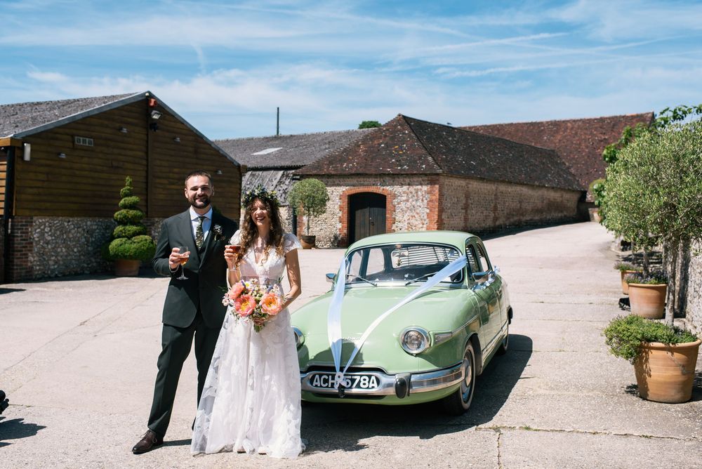 Bride and groom posing beside a vintage mint green wedding car decorated with ribbon outside a rustic barn venue.