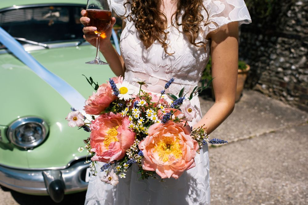 Bride in lace dress holding coral peony bouquet and champagne glass beside a mint green vintage wedding car.