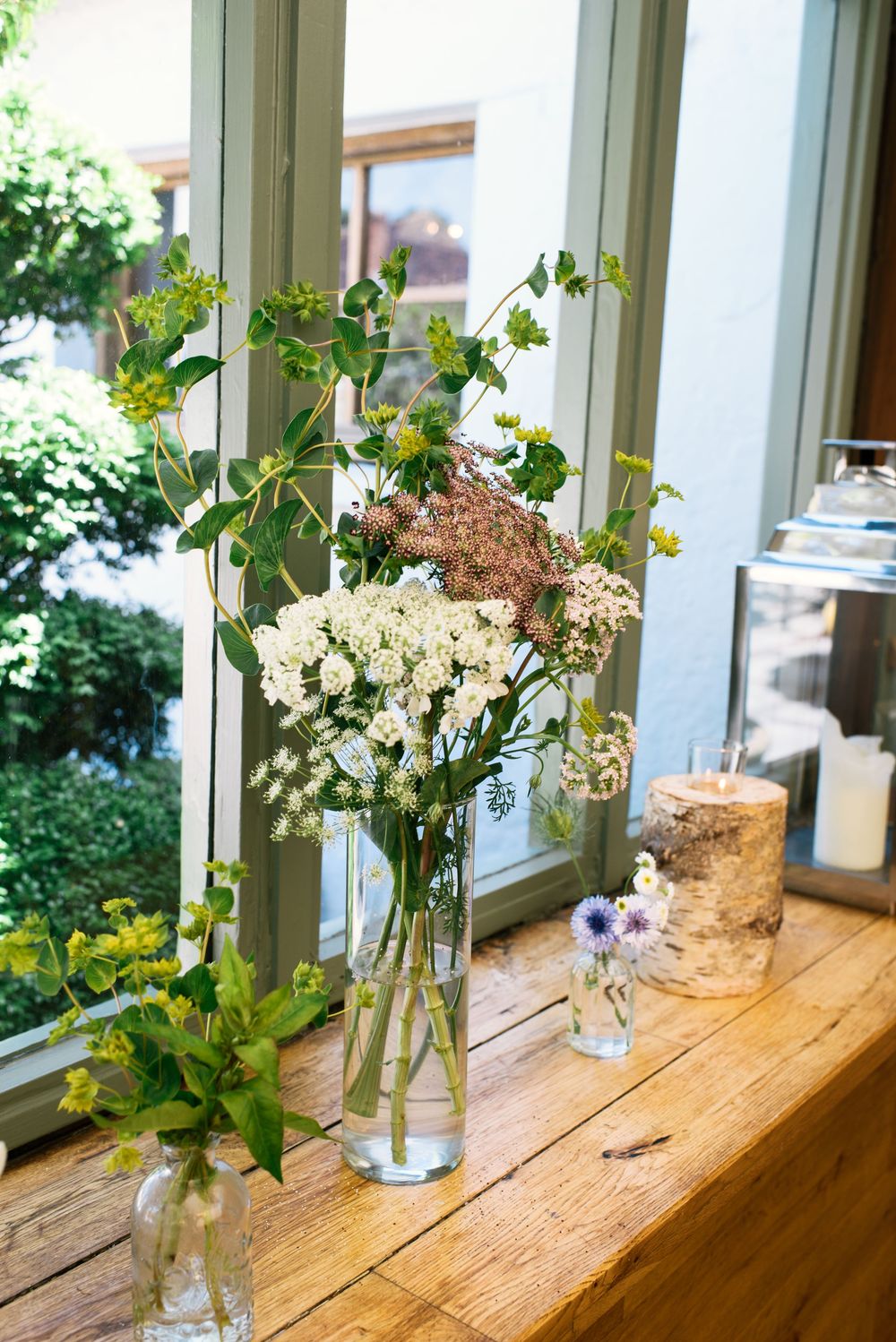 Glass vases with wildflowers and candles on a rustic wooden windowsill with green-framed windows.