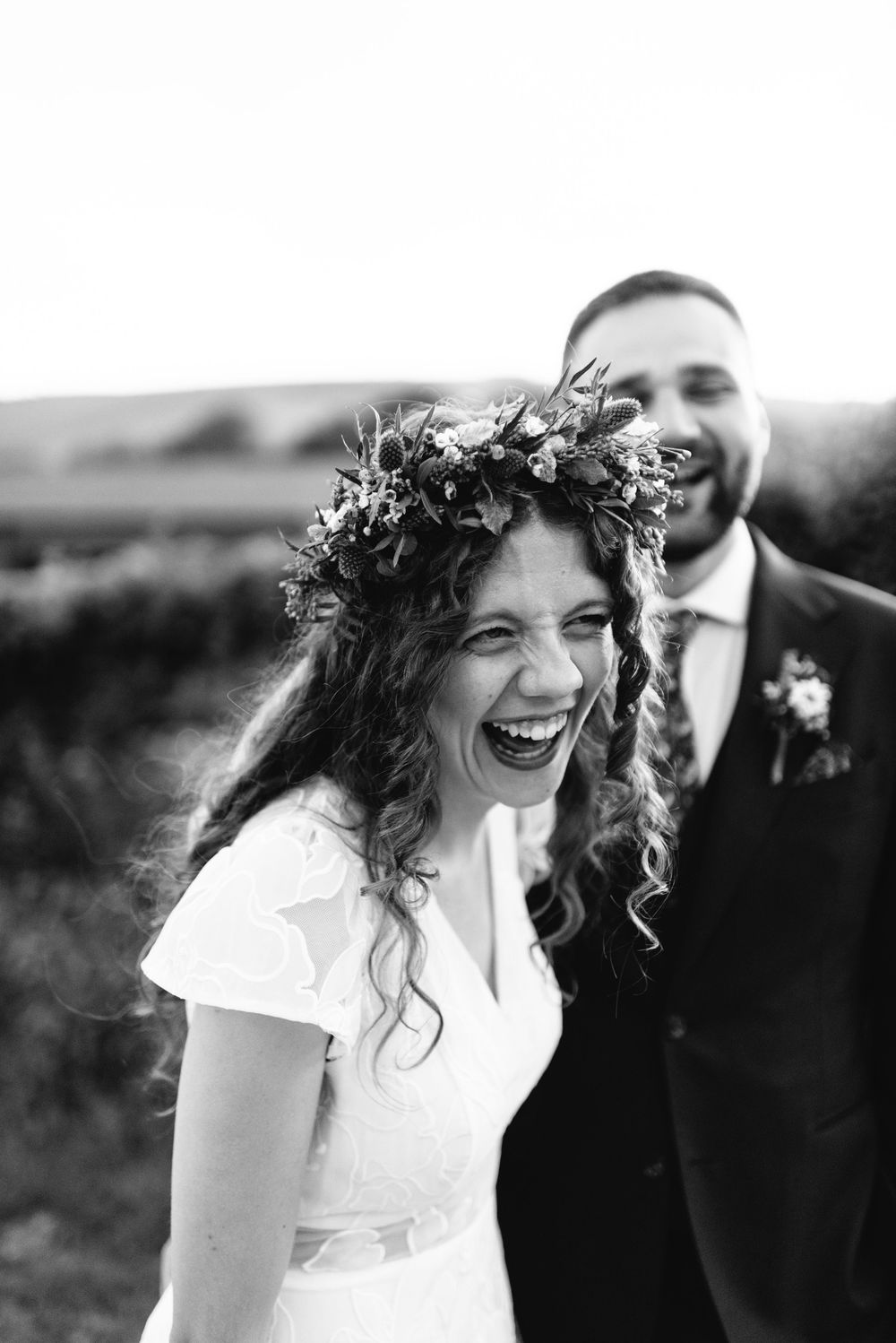 Black and white photo of a laughing bride wearing a floral crown with groom smiling behind her at outdoor wedding.