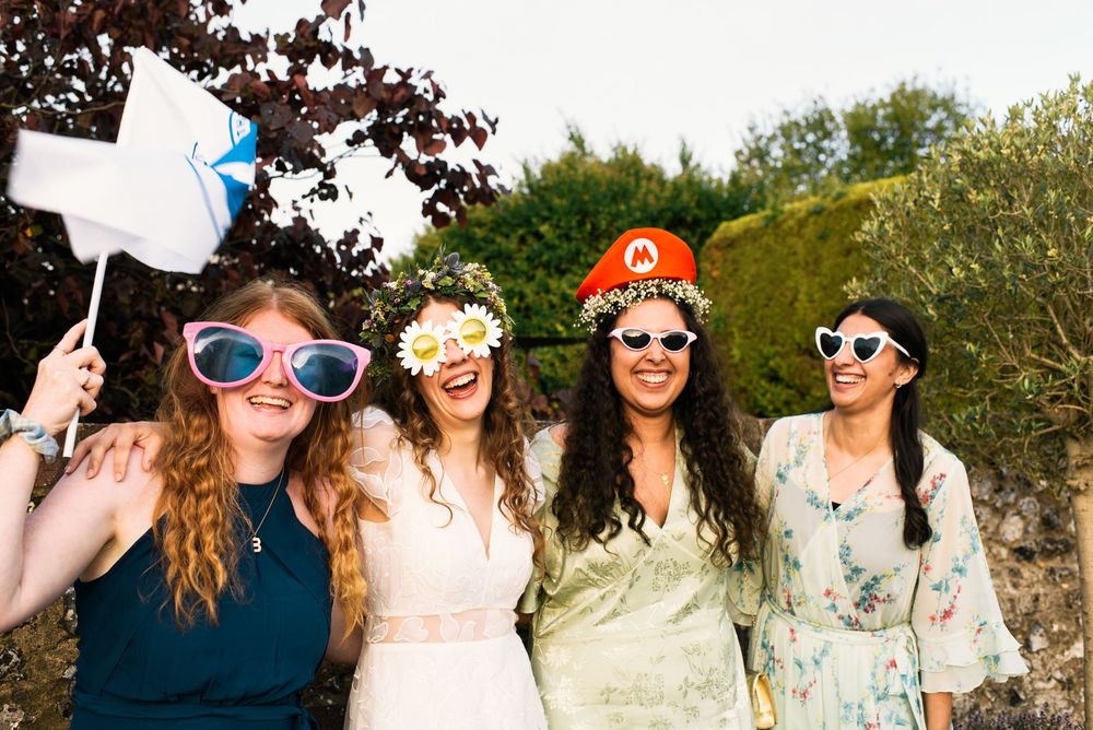 Four women laughing outdoors wearing fun accessories including flower crowns, Mario hat, and oversized sunglasses at a party.