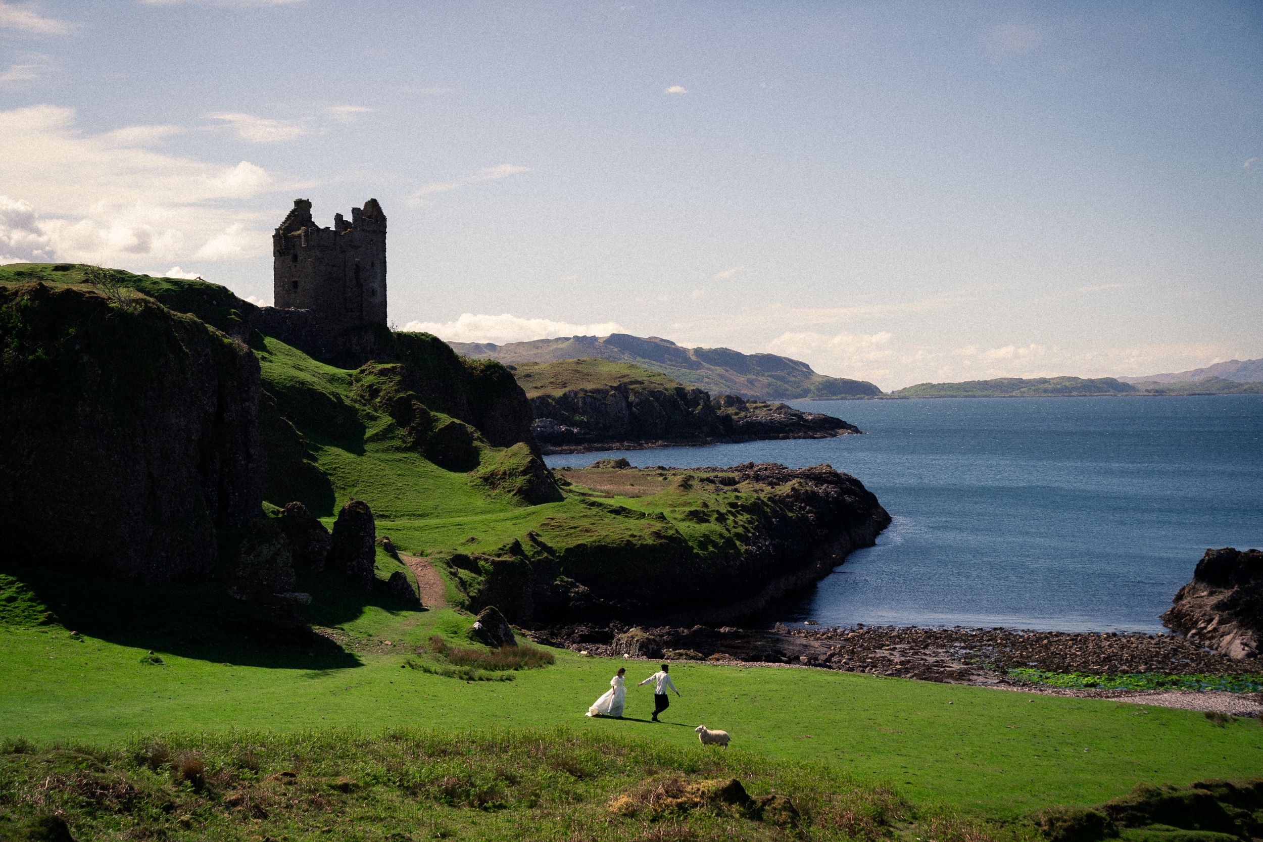 Couple walking toward ancient Scottish castle on green coastal cliffs with scenic loch and mountains beyond.