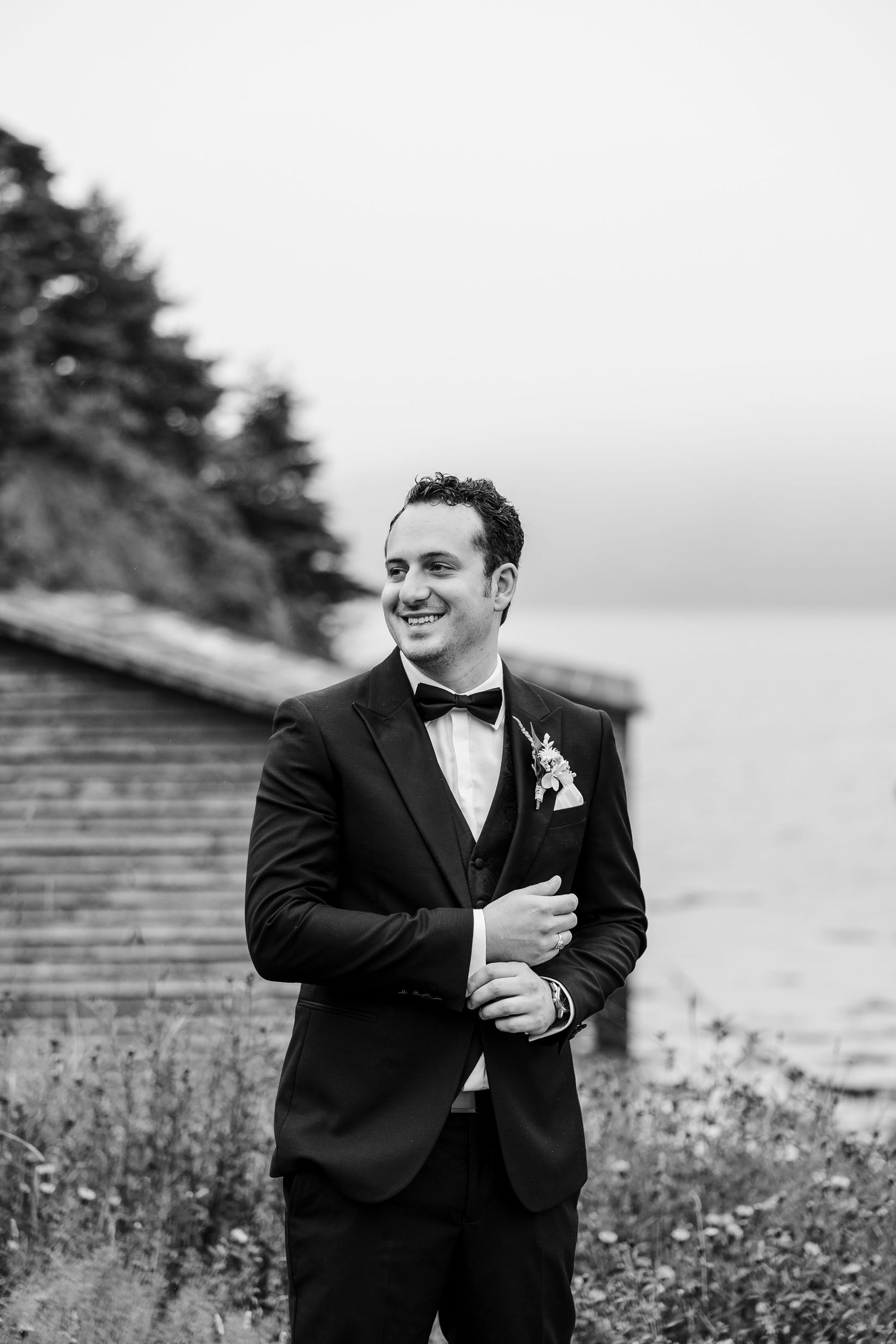Smiling groom in black tuxedo with boutonniere poses outdoors near waterfront in elegant black and white wedding photo.
